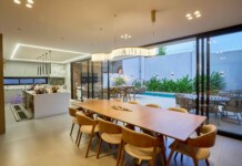 Open-concept kitchen and dining area with a long wooden table, beige chairs, pendant lights, and glass doors to a pool patio outside.