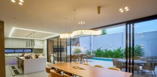 Open-concept kitchen and dining area with a long wooden table, beige chairs, pendant lights, and glass doors to a pool patio outside.
