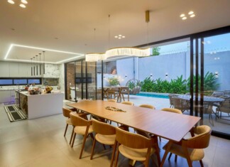 Open-concept kitchen and dining area with a long wooden table, beige chairs, pendant lights, and glass doors to a pool patio outside.