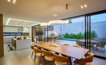 Open-concept kitchen and dining area with a long wooden table, beige chairs, pendant lights, and glass doors to a pool patio outside.