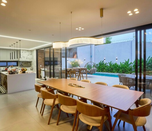 Open-concept kitchen and dining area with a long wooden table, beige chairs, pendant lights, and glass doors to a pool patio outside.