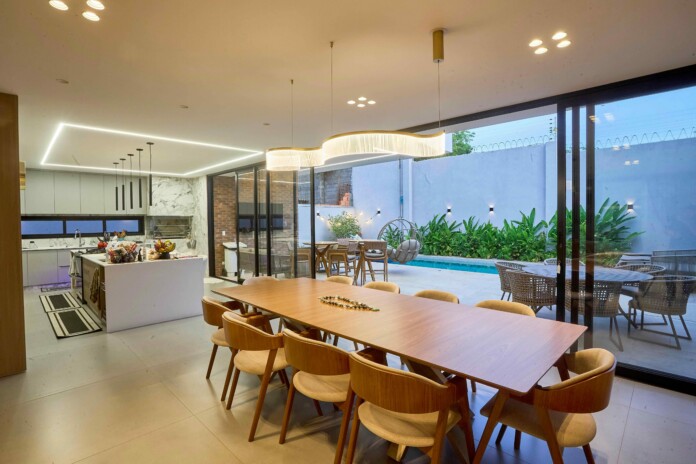 Open-concept kitchen and dining area with a long wooden table, beige chairs, pendant lights, and glass doors to a pool patio outside.