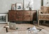 Cozy living room scene with a wooden sideboard, wicker chair, laptop open on a rug, and scattered magazines and an open notebook in the foreground