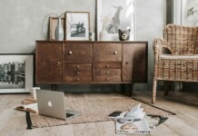 Cozy living room scene with a wooden sideboard, wicker chair, laptop open on a rug, and scattered magazines and an open notebook in the foreground