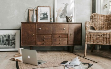 Cozy living room scene with a wooden sideboard, wicker chair, laptop open on a rug, and scattered magazines and an open notebook in the foreground