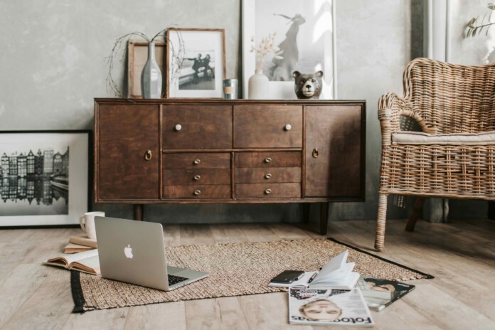 Cozy living room scene with a wooden sideboard, wicker chair, laptop open on a rug, and scattered magazines and an open notebook in the foreground