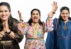 Three smiling women of Indian descent raise their hands in celebration against a white background, wearing colorful outfits and jewelry.