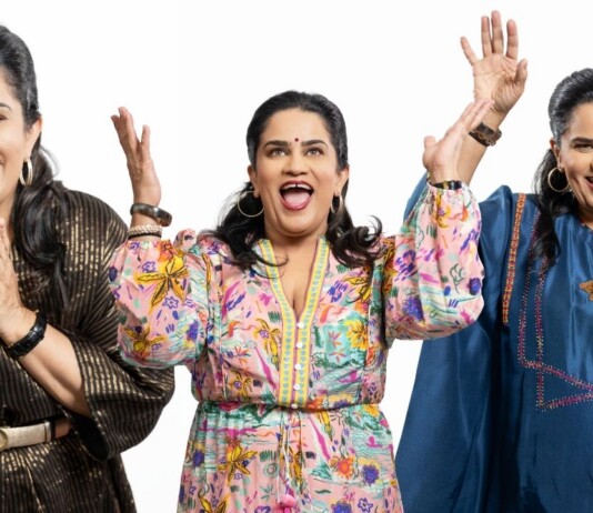 Three smiling women of Indian descent raise their hands in celebration against a white background, wearing colorful outfits and jewelry.