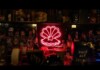 Neon pink seashell sign with a pearly center glows behind a bar, surrounded by assorted liquor bottles.