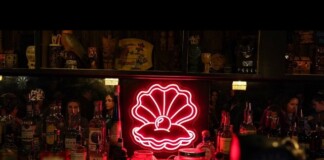 Neon pink seashell sign with a pearly center glows behind a bar, surrounded by assorted liquor bottles.