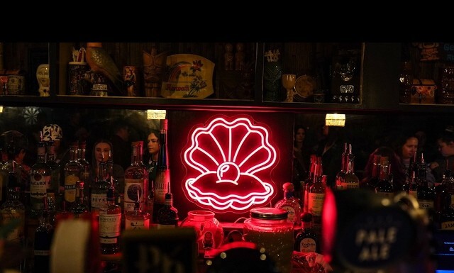 Community Rallies to Save Denver’s Only Lesbian Bar With $80K Lifeline Neon pink seashell sign with a pearly center glows behind a bar, surrounded by assorted liquor bottles.