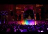 Speaker at a podium on a rainbow-lit stage in a grand, dark hall, audience seated at tables below.
