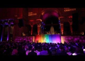 Speaker at a podium on a rainbow-lit stage in a grand, dark hall, audience seated at tables below.