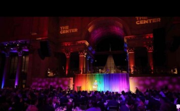 Speaker at a podium on a rainbow-lit stage in a grand, dark hall, audience seated at tables below.