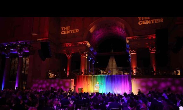 Speaker at a podium on a rainbow-lit stage in a grand, dark hall, audience seated at tables below.