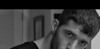 Close-up black-and-white portrait of a man with short hair and beard, gazing intently at the camera with a serious expression.