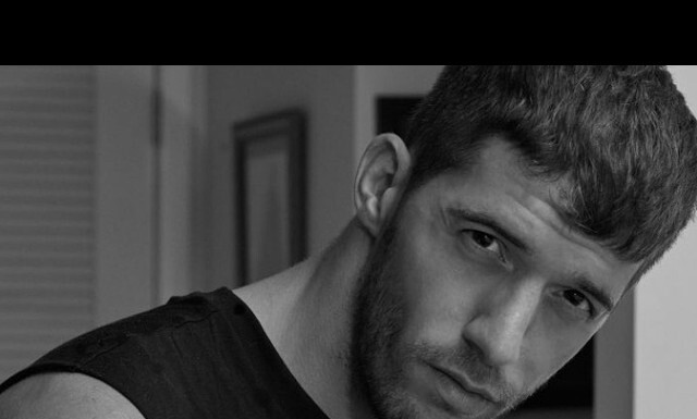 Close-up black-and-white portrait of a man with short hair and beard, gazing intently at the camera with a serious expression.