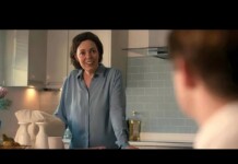 Woman in a blue blouse leans on a kitchen counter, smiling at a person whose back is visible in a modern tiled kitchen.