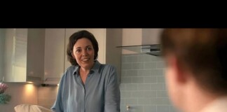 Woman in a blue blouse leans on a kitchen counter, smiling at a person whose back is visible in a modern tiled kitchen.