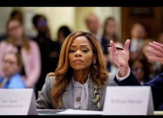 Panelist in a gray blazer with a decorative brooch sits at a table, facing an audience.
