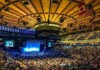 Large indoor arena full of seated attendees watching a blue-lit stage perform.