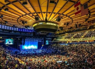 Large indoor arena full of seated attendees watching a blue-lit stage perform.