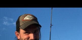 Smiling man wearing a baseball cap holds a fishing rod in a clear blue sky selfie.