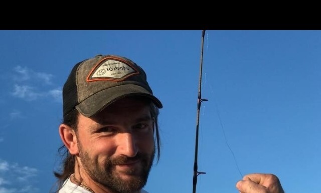 Smiling man wearing a baseball cap holds a fishing rod in a clear blue sky selfie.