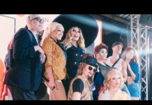 Group of people posing on a stage with bright lights and metal truss, wearing fashionable outfits including a sequined jacket and hats.