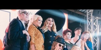 Group of people posing on a stage with bright lights and metal truss, wearing fashionable outfits including a sequined jacket and hats.