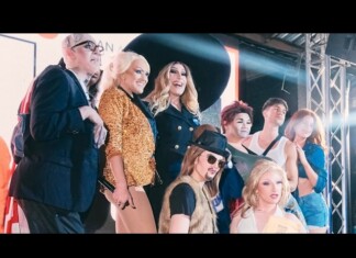 Group of people posing on a stage with bright lights and metal truss, wearing fashionable outfits including a sequined jacket and hats.