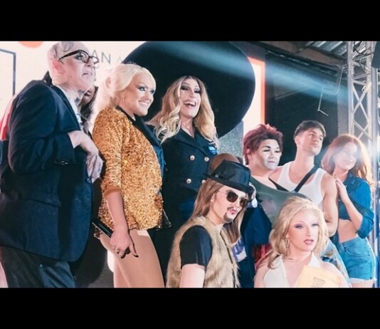 Group of people posing on a stage with bright lights and metal truss, wearing fashionable outfits including a sequined jacket and hats.