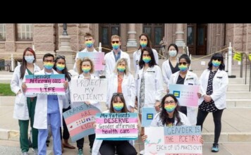 Group of students in white coats with rainbow masks holding signs advocating gender-affirming care and LGBTQ rights on courthouse steps.