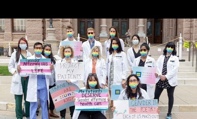 Group of students in white coats with rainbow masks holding signs advocating gender-affirming care and LGBTQ rights on courthouse steps.