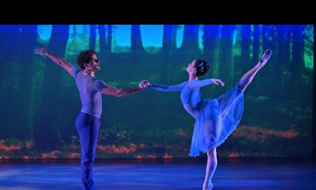 Two ballet dancers perform a duet on a blue-lit stage, with a forest backdrop; the man on the left holds the woman's hand as she balances en pointe in an arabesque.