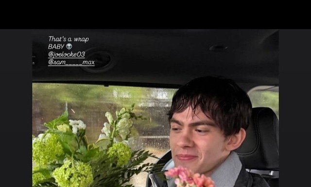 Young man in a car, smiling with pink and green flowers in the foreground; text overlay reads 'That's a wrap BABY' with social handles (joelocke03, sam__max).