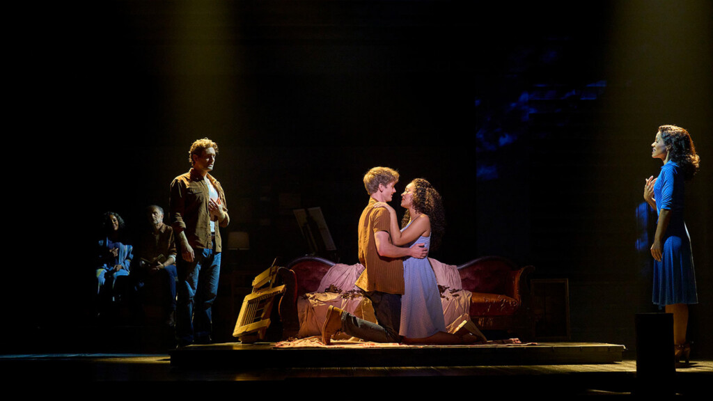 The Notebook’s Broadway Touring Company – Comes to the Kravis and Arsht Centers | Hotspots Magazine Hotspots Magazine Theatrical stage scene: a couple kneels on a bed and embraces while several other actors stand or sit off to the side under warm lighting.