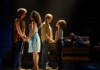 Four actors stand in two couples on a dimly lit stage, holding hands as if in rehearsal, with a couple seated on a sofa in the background.