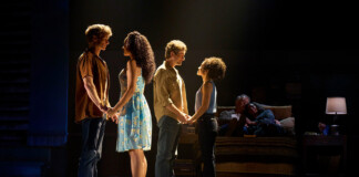 Four actors stand in two couples on a dimly lit stage, holding hands as if in rehearsal, with a couple seated on a sofa in the background.