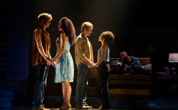 Four actors stand in two couples on a dimly lit stage, holding hands as if in rehearsal, with a couple seated on a sofa in the background.