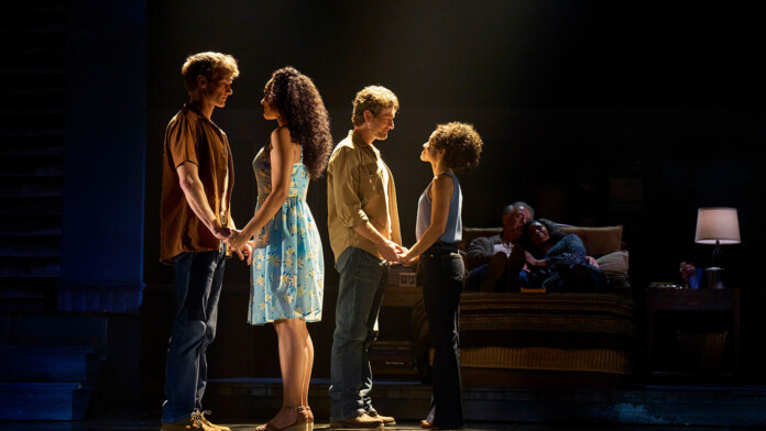 Four actors stand in two couples on a dimly lit stage, holding hands as if in rehearsal, with a couple seated on a sofa in the background.