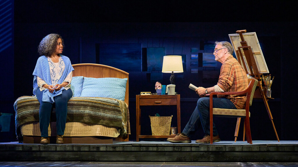 Two actors sit across from each other on a bedroom set: a woman on the bed facing a man in a chair, with a nightstand, lamp, and easel nearby.