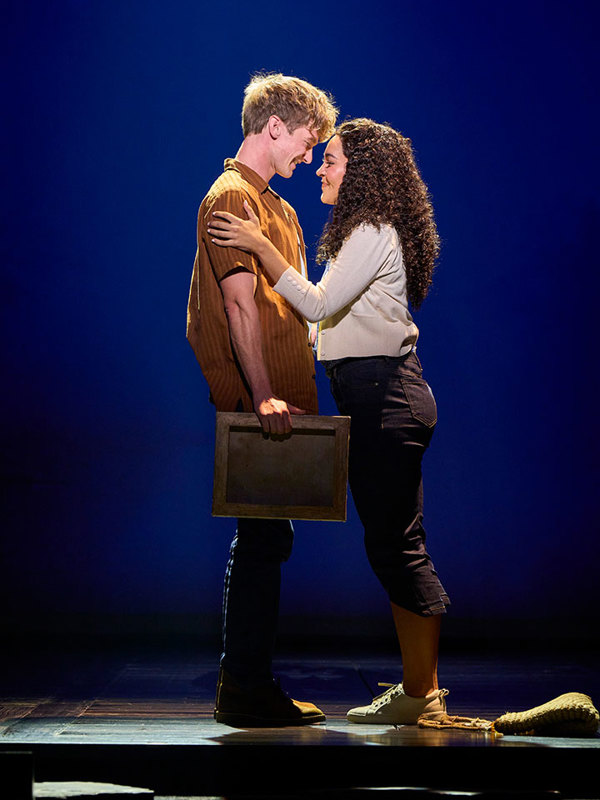 Two people stand close on a stage, facing and touching foreheads, one holding a wooden frame, under blue spotlight.