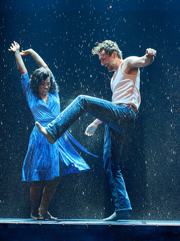 A man in a white tank top and jeans and a woman in a blue dress dancing joyfully in the rain on a stage.