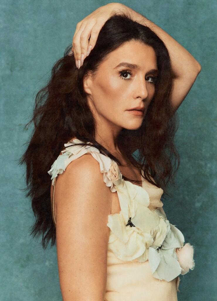 Portrait of a woman with long dark hair, hand resting on her head, wearing a cream ruffled dress against a teal textured backdrop.