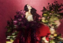 Woman with long dark hair in a strapless white top, posing among vibrant pink flowers against a pink backdrop.
