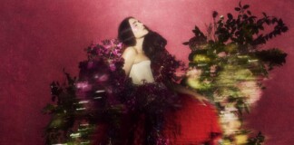 Woman with long dark hair in a strapless white top, posing among vibrant pink flowers against a pink backdrop.