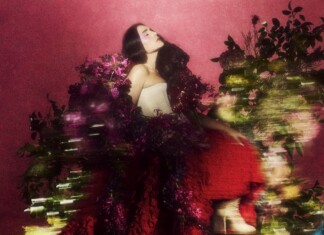 Goddess in Sneakers – Jessie Ware on bigger vocals, queer fans who lift her up and the joy of wigs Woman with long dark hair in a strapless white top, posing among vibrant pink flowers against a pink backdrop.