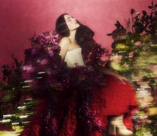 Woman with long dark hair in a strapless white top, posing among vibrant pink flowers against a pink backdrop.