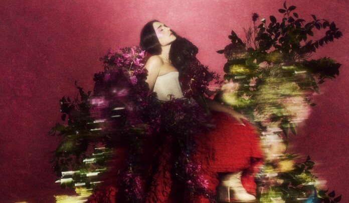 Woman with long dark hair in a strapless white top, posing among vibrant pink flowers against a pink backdrop.